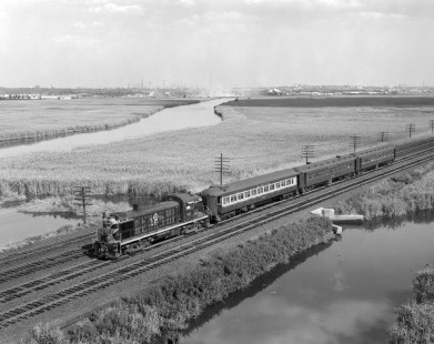Erie Lackawanna Railroad no. 921 leads a Suffern-bound passenger train through Rutherford, New Jersey, on July 20, 1965. The Manhattan skyline and the Empire State Building are visible on the horizon. Photograph by Victor Hand. Hand-EL-30-062.JPG.
