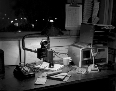 An artful still-life perspective of the interior of the Illinois Central Gulf yard office in Bloomington, Indiana, during the mid-to late-1970s.

Judges’ Comments: This photograph masterfully captures the essence of an operator’s life within the confines of an Illinois Central yard office nestled in Bloomington, Indiana. The diverse tools of the trade glow in the pool of a single lightbulb forming an intricate still-life where several generations of train-control technologies are surrounded by the marks of the operator’s stained coffee cup and creep of cigarette ash in the ashtray. These meticulous details add a layer of authenticity to the scene, including the subtle marks of weathered varnish and frayed wires adorning the operator's desk, foretelling the ever-present passage of time.

Read more about the 2023 John E. Gruber Creative Photography Awards: <a href="https://railphoto-art.org/awards-2023/" rel="noreferrer nofollow">railphoto-art.org/awards-2023/</a>
