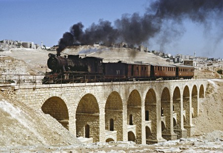 On the Hedjaz Jordan Railway, steam locomotive no. 71 leads a three-car passenger train across a stone arch viaduct, which carries an aqueduct on its lower level. The view is from the outskirts of Amman on July 15, 1991. Photograph by Fred M. Springer, © 2016, Center for Railroad Photography and Art. Springer-Hedjaz-ZimZam(1)-02-20
