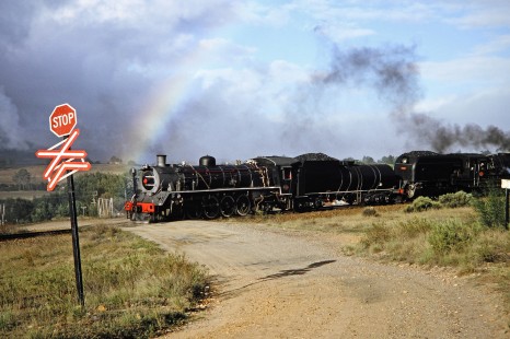 South African Railway steam locomotives nos. 2683 and 4122 move past a railroad crossing with a rainbow in the background in Camfer, Western Cape, South Africa, on March 23, 1995. Photograph by Fred M. Springer, © 2014, Center for Railroad Photography and Art. Springer-So.Africa(1)-16-01