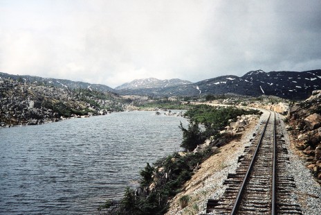 White Pass & Yukon Railroad tracks by lake waters and craggy mountains in Bennett, British Columbia, Canada, on June 13, 1998. Photograph by Fred M. Springer, © 2014, Center for Railroad Photography and Art. Springer-Alaska-NZ-10-13