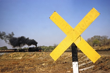 A National Railways of Zimbabwe Garratt steam locomotive heads toward a "yellowed" out railroad crossing sign in Hwange, Matabeleland, Zimbabwe, on August 6, 1991. Photograph by Fred M. Springer, © 2014, Center for Railroad Photography and Art. Springer-Hedjaz-ZimZam(1)-24-27