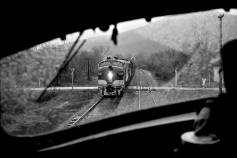 The windshield of a northbound Baltimore & Ohio freight train frames a southbound train at rural crossing just north of Bradford, Pennsylvania, on the B&O’s former Buffalo, Rochester & Pittsburg line on a rainy May 13th, 1968. The southbound is using power borrowed from the Chesapeake & Ohio, showing influence that would eventually lead to the “Chessie System” mergers. Note the photographer at far right: Ken's wife, Patricia Kraemer, photographing her husband's train.

Read more about the <a href="http://www.railphoto-art.org/awards/2017-awards/" rel="nofollow">2017 John E. Gruber Creative Photography Awards Program</a>.