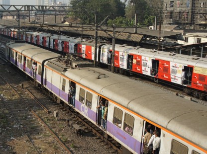 Two departing (wide gauge trains designed to hold more passengers) trains on parallel tracks gather speed a mile north of Victoria Station in Mumbai on a very hot and humid late afternoon, as passengers stand by open doors for cooler air in the non air-conditioned coaches, in 2011. © Todd Halamka