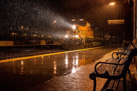 A sudden rain drenching the station platform and bench combined with a westbound manifest running on the eastward main with headlights illuminating the raindrops provide the elements for this image.